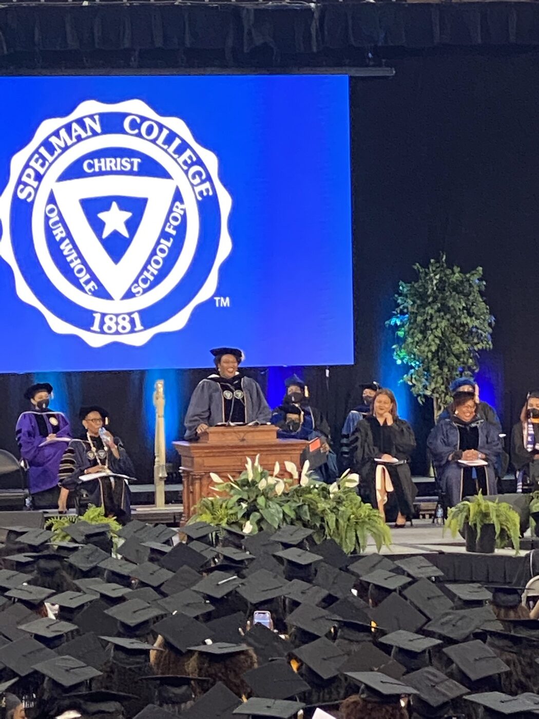 Spelman College commencement ceremony with rows of graduates in caps and gowns, the Spelman seal displayed on stage