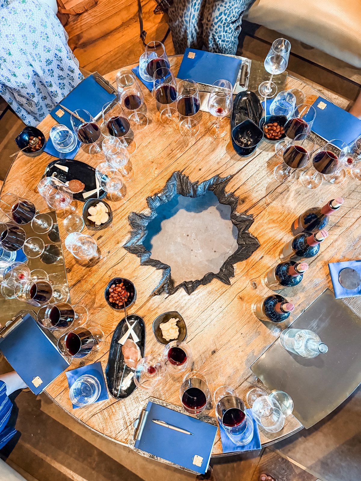Overhead view of the tasting table at Covert Estate in Napa, with wine glasses, pours, blue notebooks, and small plates arranged around the table’s open center.